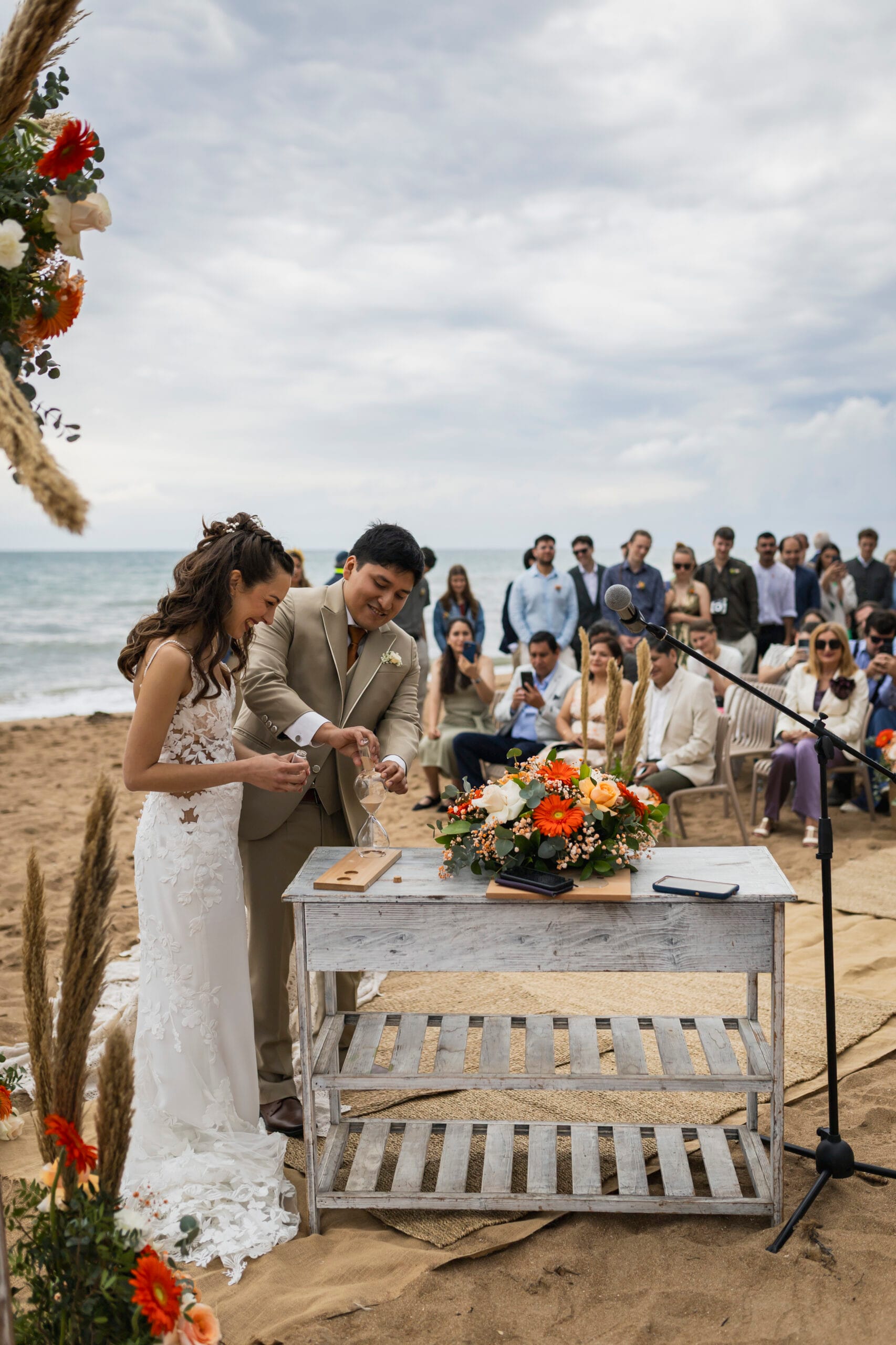 Novios sonrientes realizando el ritual de la arena en la playa durante una boda organizada por Restaurante Escondida frente al mar.