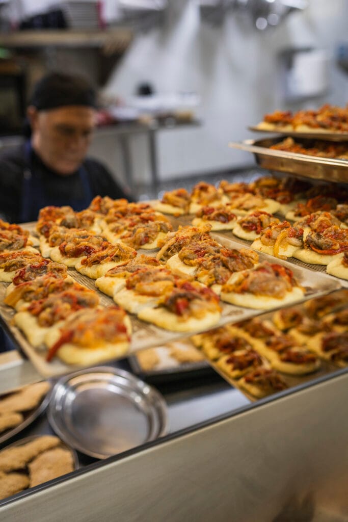 Bandejas repletas de cocas de verduras tradicionales recién horneadas listas para el servicio de catering, con un chef trabajando de fondo en las cocinas de Restaurante Escondida.